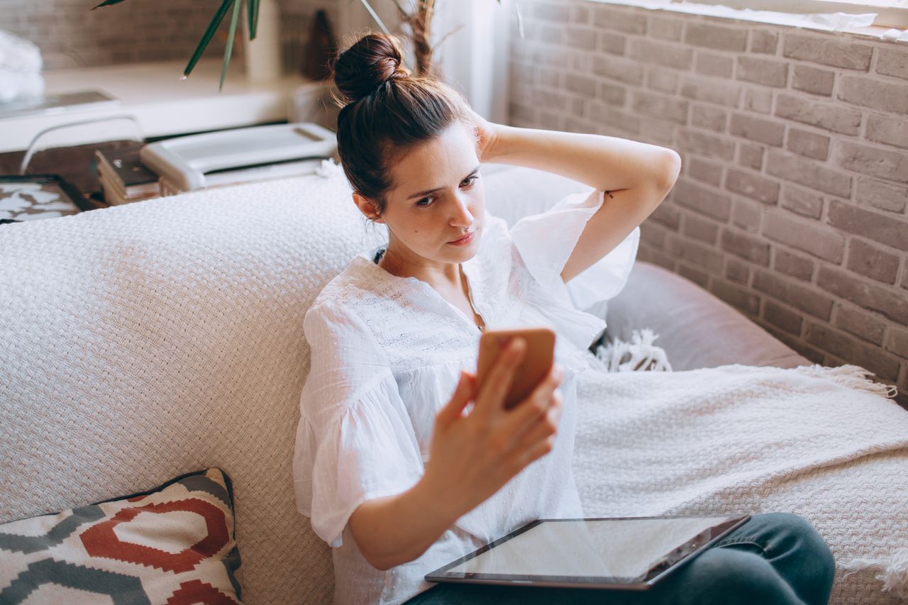 services-01 A woman adjusts her hair while taking a selfie on a smartphone, seated on a sofa with a tablet, embodying remote work lifestyle.